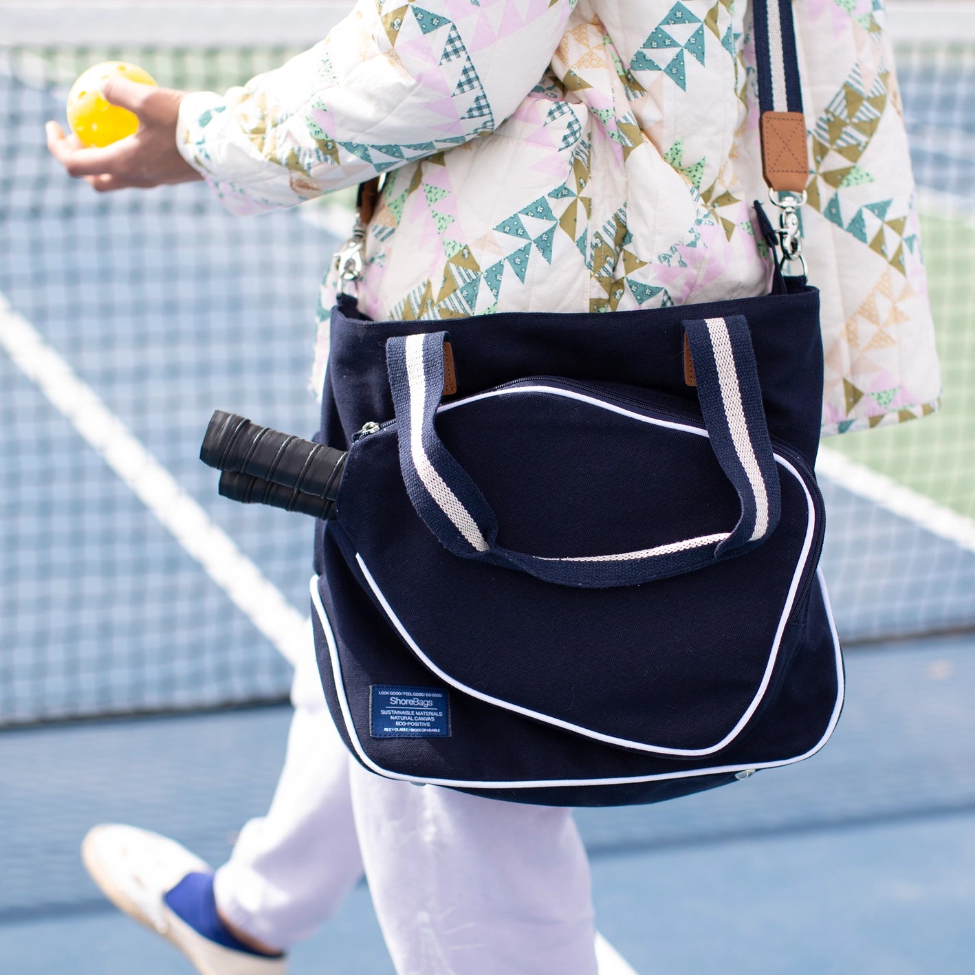 Person on a pickleball court with a sleek navy blue pickleball bag hanging over their shoulder with two paddles in it and a ball in their hand.