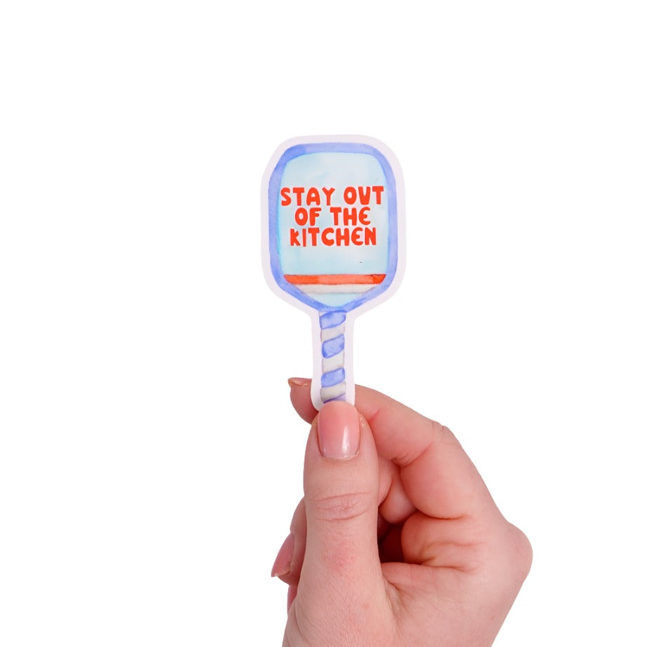 Blue sticker shaped like a pickleball paddle that says "STAY OUT OF THE KITCHEN" on it in red being held