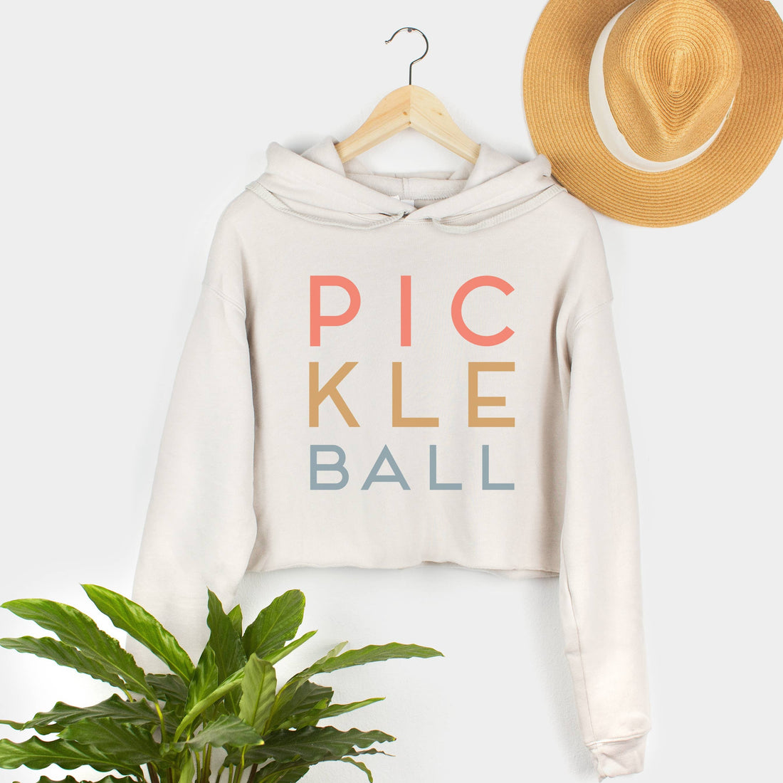 Hanging cream colored cropped hoodie sweatshirt that says "PICKLEBALL" on it in colorful lettering with a straw hat hanging above it and a plant sitting below it