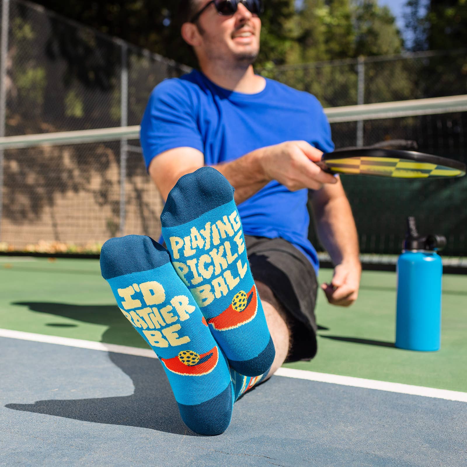 Man sitting on a pickleball court wearing humorous blue socks that say "I"D RATHER BE PLAYING PICKLEBALL" on the soles of them