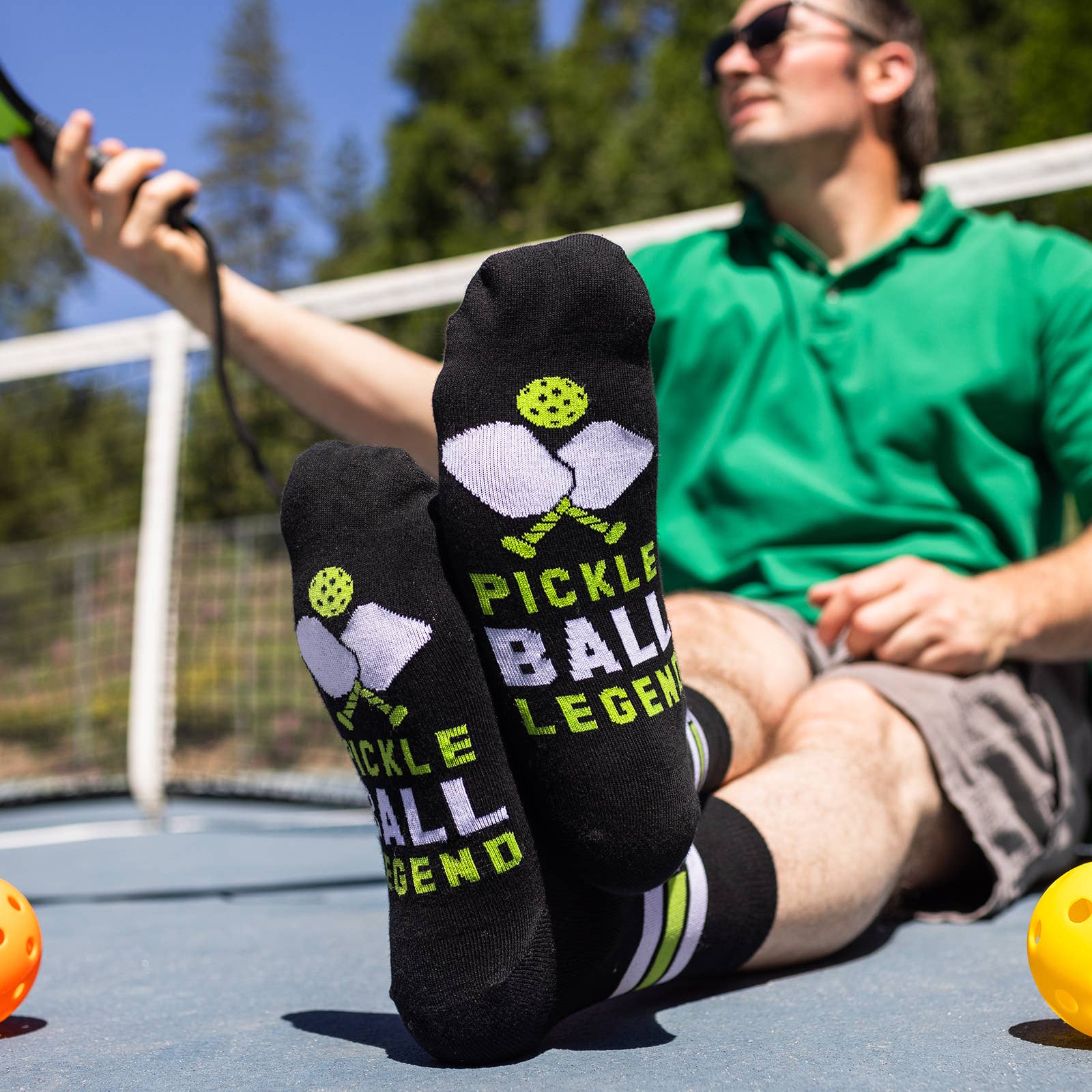 Man sitting on a pickleball court leaning against the net with black socks on that say Pickleball Legend on the bottom of the socks