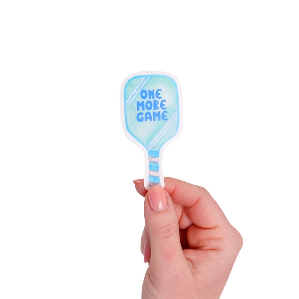 Blue sticker shaped like a pickleball paddle that says "ONE MORE GAME" on it being held