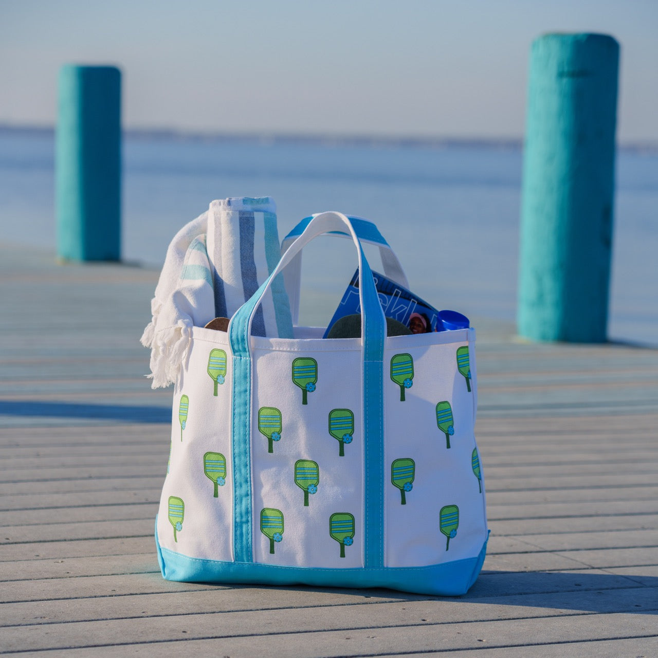 Canvas beach tote with a green pickleball paddle print on it and blue handles filled with a beach towel and magazine sitting on a pier with water in the background