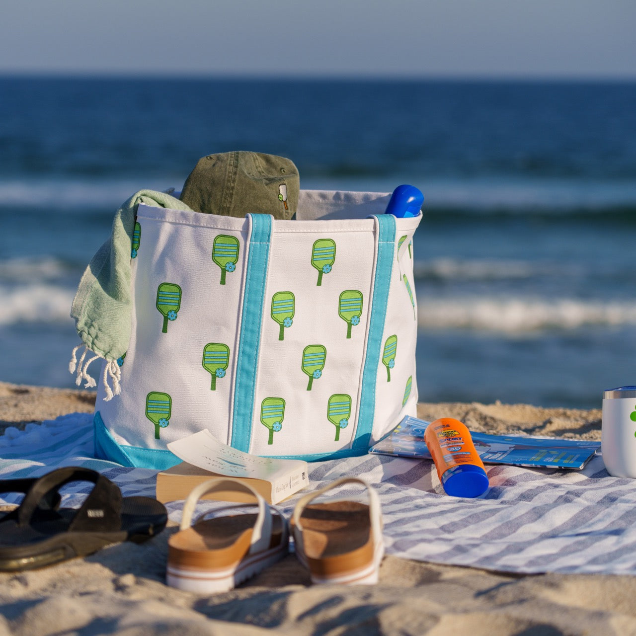 Canvas beach tote with a green pickleball paddle print on it and blue handles filled with a beach towel and magazine sitting on a beach with the ocean in the background