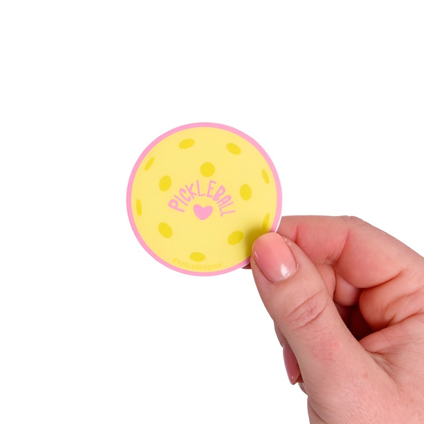 Yellow sticker shaped like a pickleball with a pink heart and pink "PICKLEBALL" on it being held