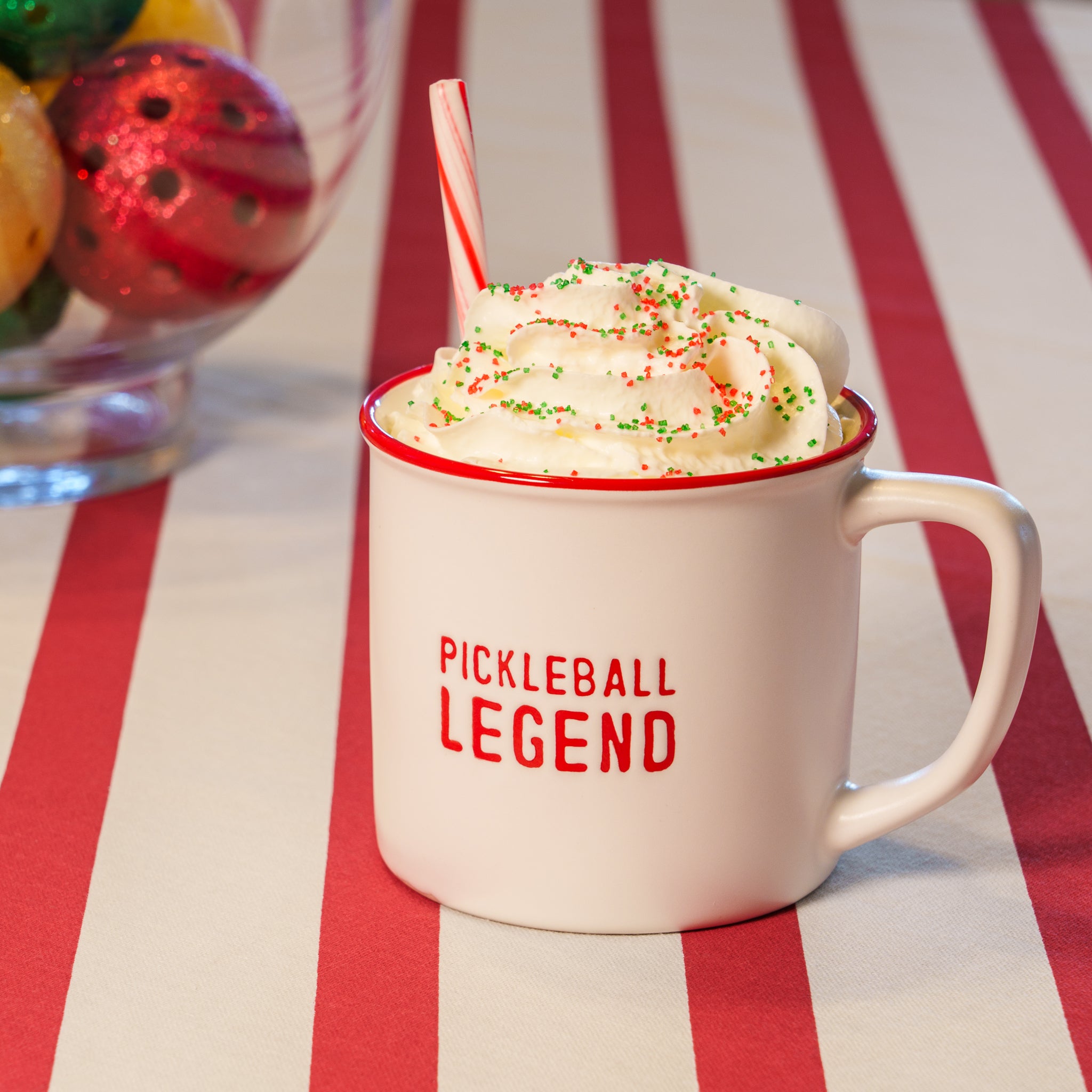 A white debossed mug that says "PICKLEBALL LEGEND" on it in red lettering with whipped cream and a candy cane in it sitting on a red and white striped table cloth