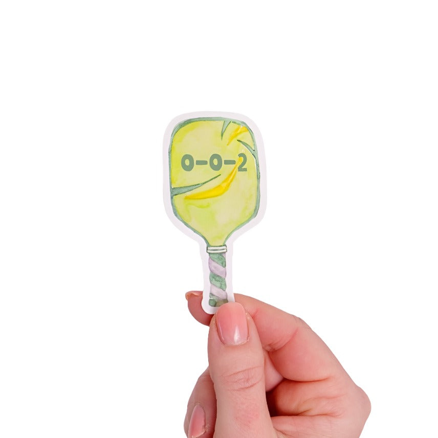 Green sticker shaped like a pickleball paddle with "0-0-2" on it being held