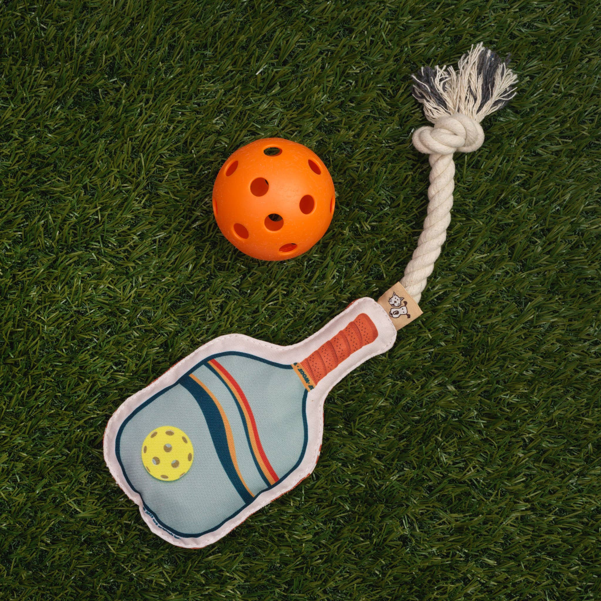 Dog toy shaped like a pickleball paddle with a rope attached sitting in grass next to an orange pickleball