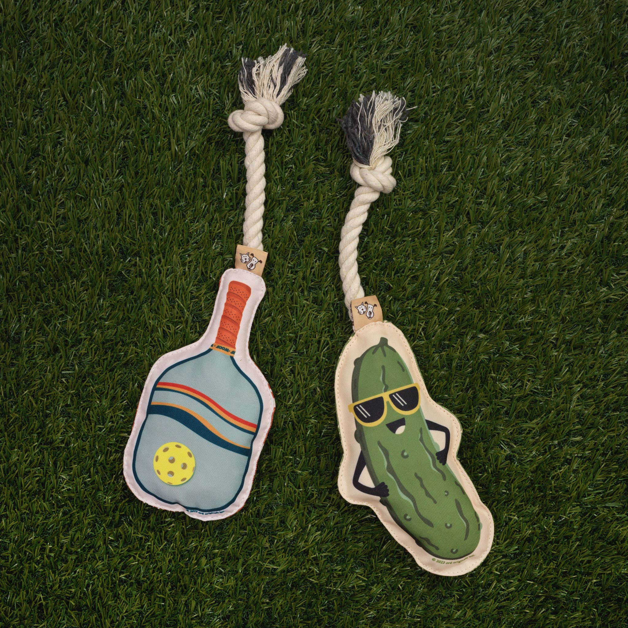 A rope dog toy shaped like a pickleball paddle and a rope dog toy shaped like a pickle laying in grass