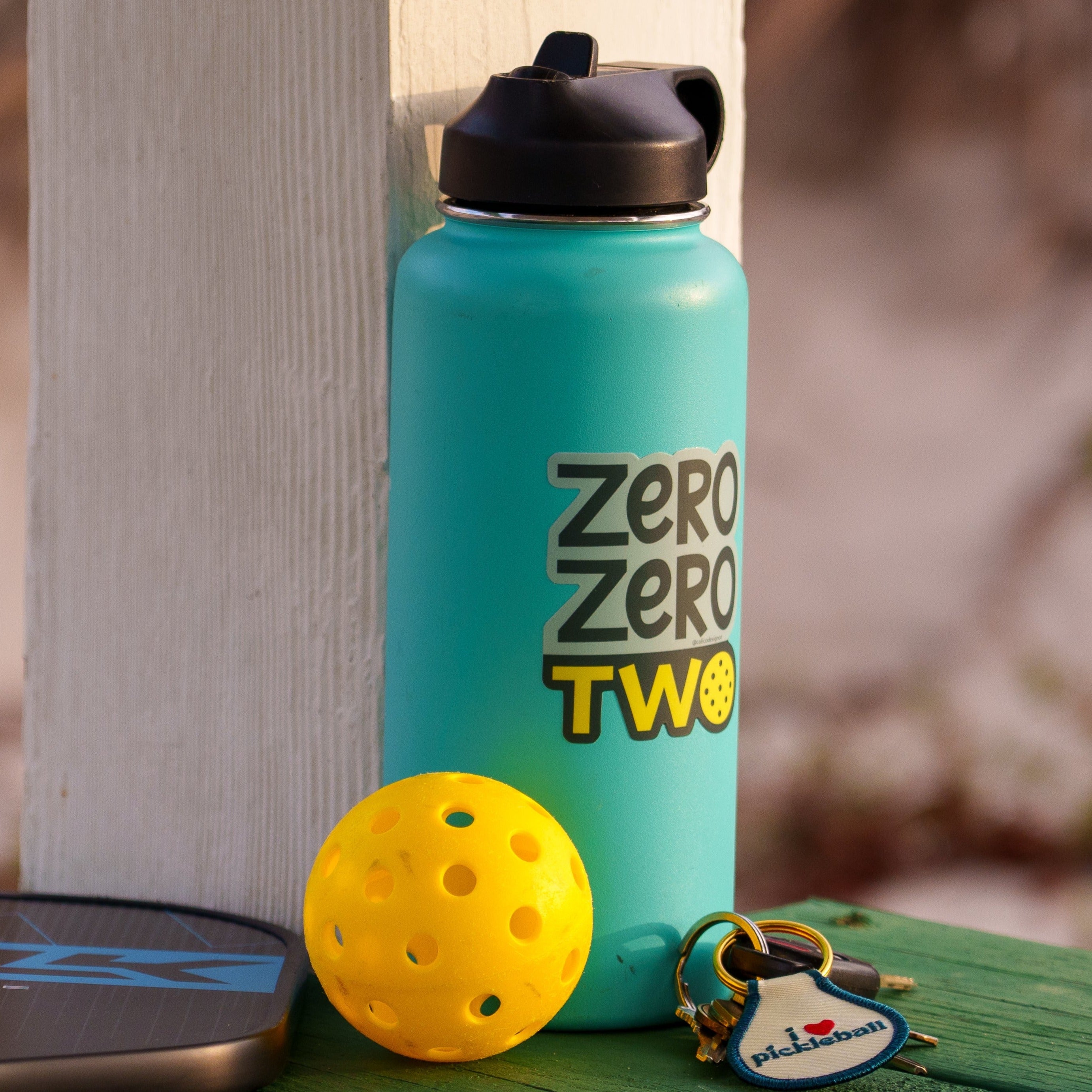 Blue water bottle with a pickleball-themed sticker on it that says "ZERO ZERO TWO" next to a pickleball paddle and ball