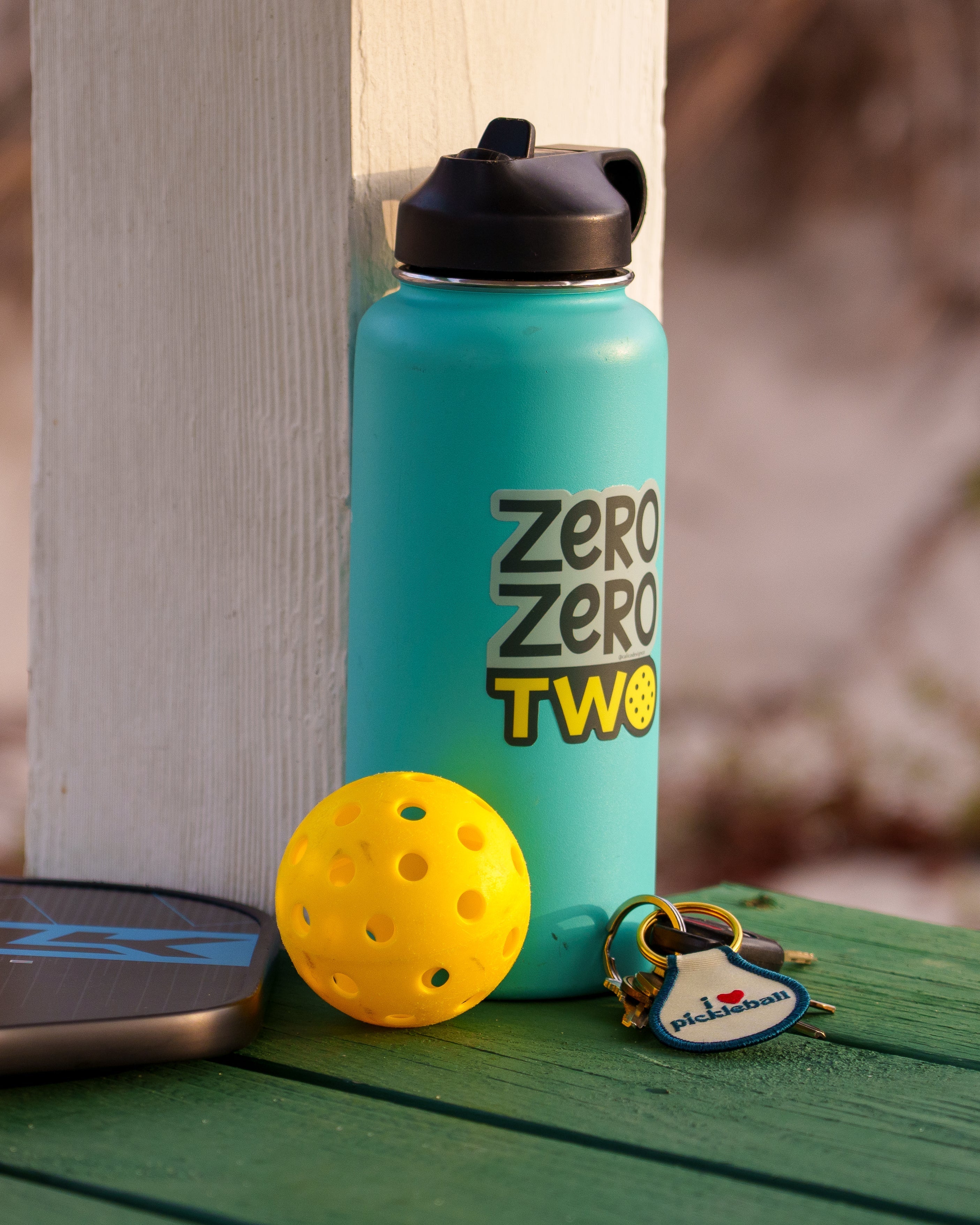 Blue water bottle with a pickleball-themed sticker on it that says "ZERO ZERO TWO" next to a pickleball paddle and ball on a boardwalk