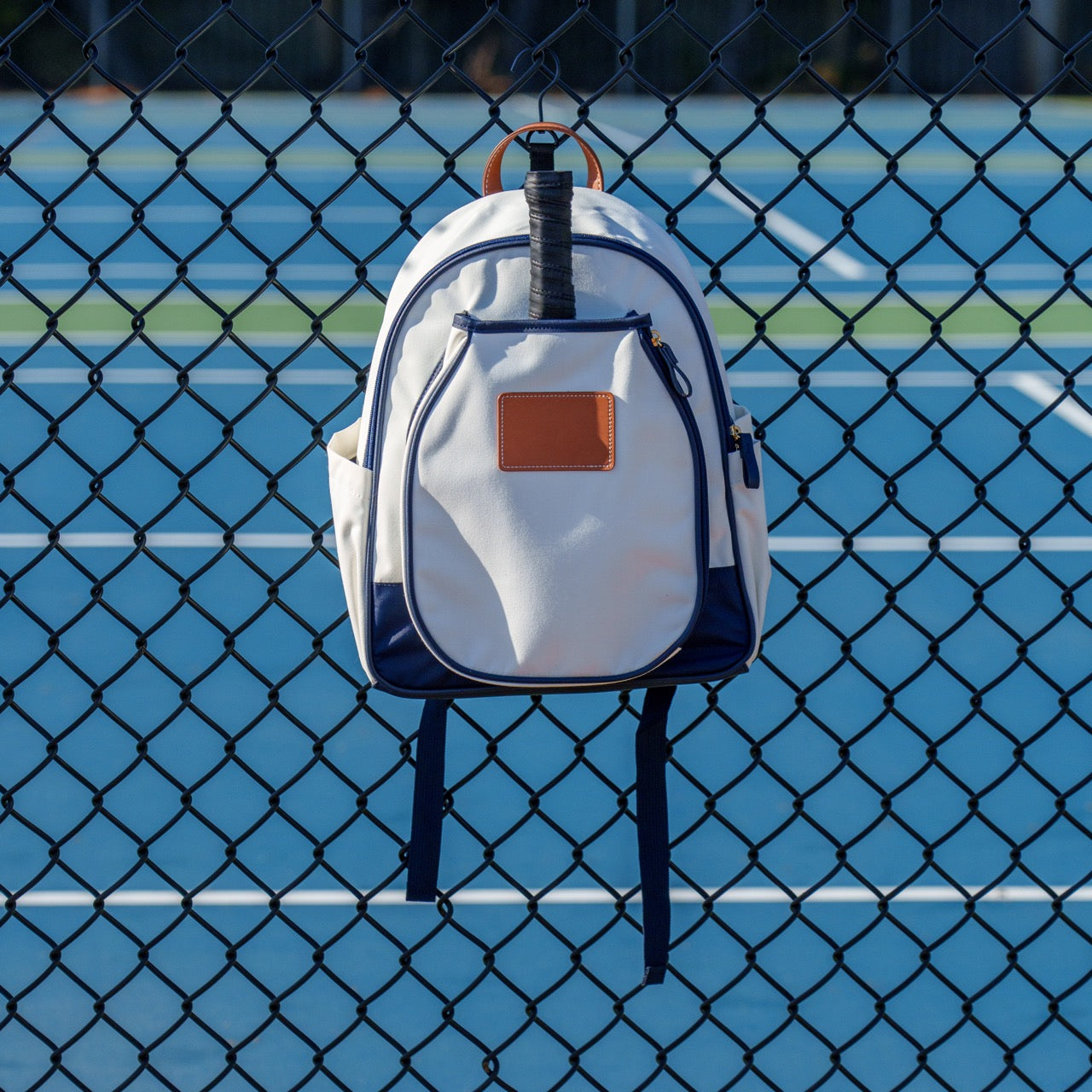 A white pickleball backpack with a pickleball paddle in it hanging on a fence in front of a pickleball court