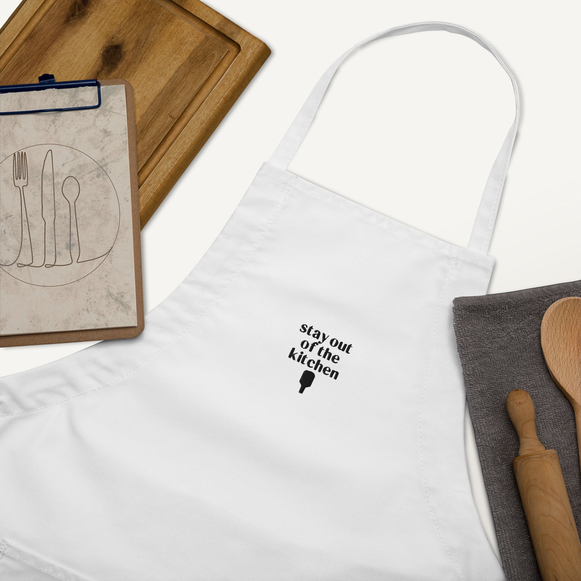 Detailed view of embroidered white apron that says "stay out of the kitchen" and has a pickleball paddle on it with a cutting board, rolling pin and wooden spoon next to it