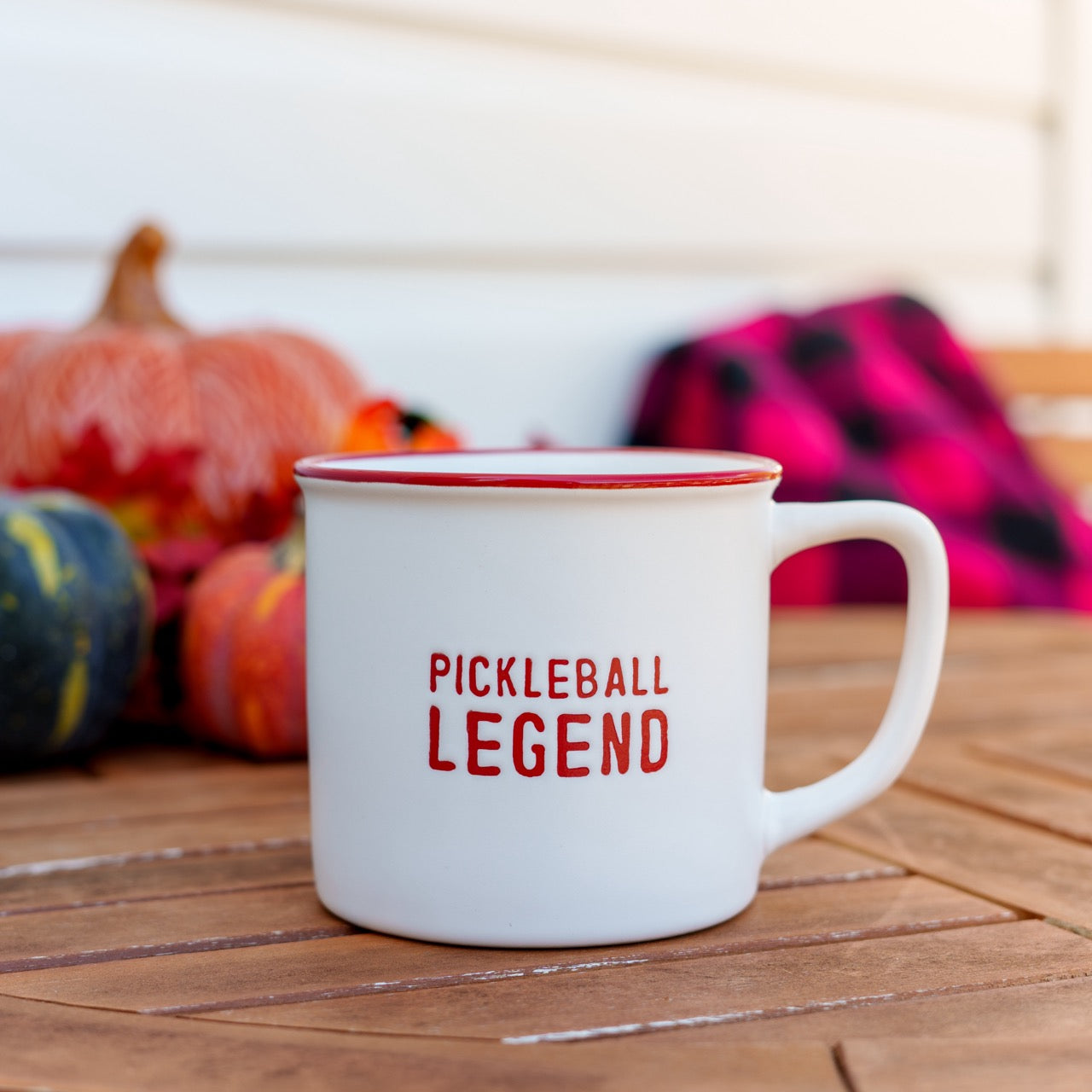White and red ceramic coffee mug that says Pickleball Legend on a table in front of pumpkins and a blanket