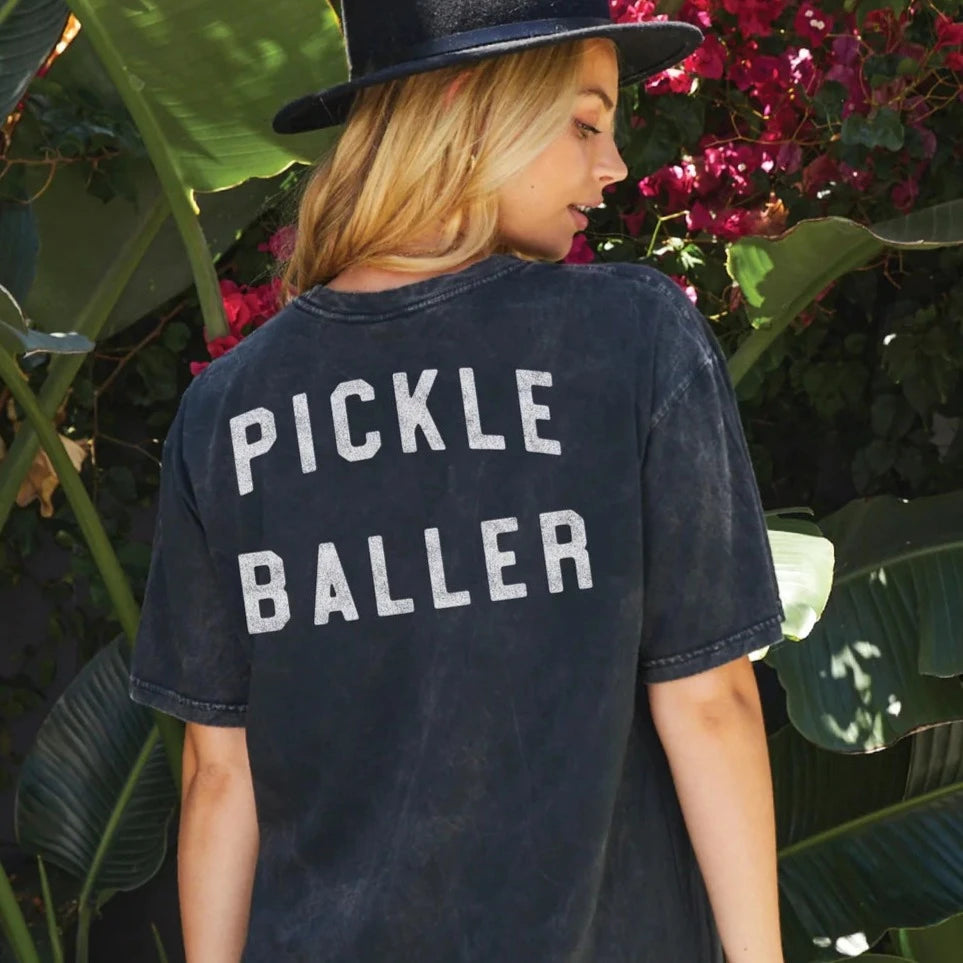 A woman wearing a washed black t-shirt that says "PICKLEBALLER" on the back and a wide brimmed black hat