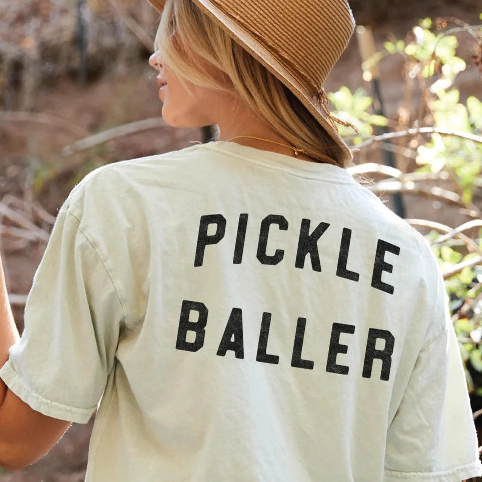 A woman wearing a cream t-shirt that says "PICKLEBALLER" on the back and a wide brimmed straw hat