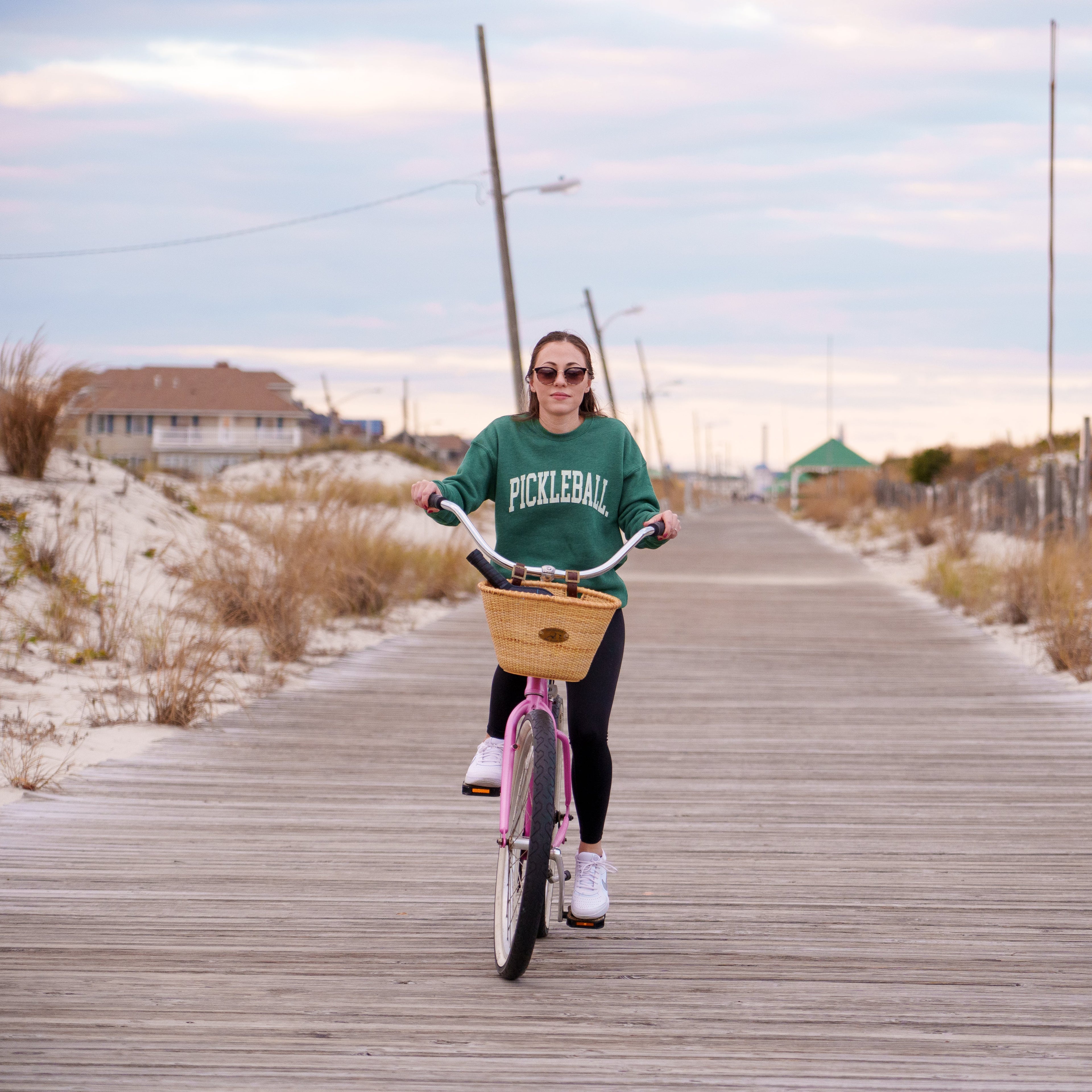 Woman riding a bike on a boardwalk wearing a green crew neck sweatshirt with the text PICKLEBALL on the front