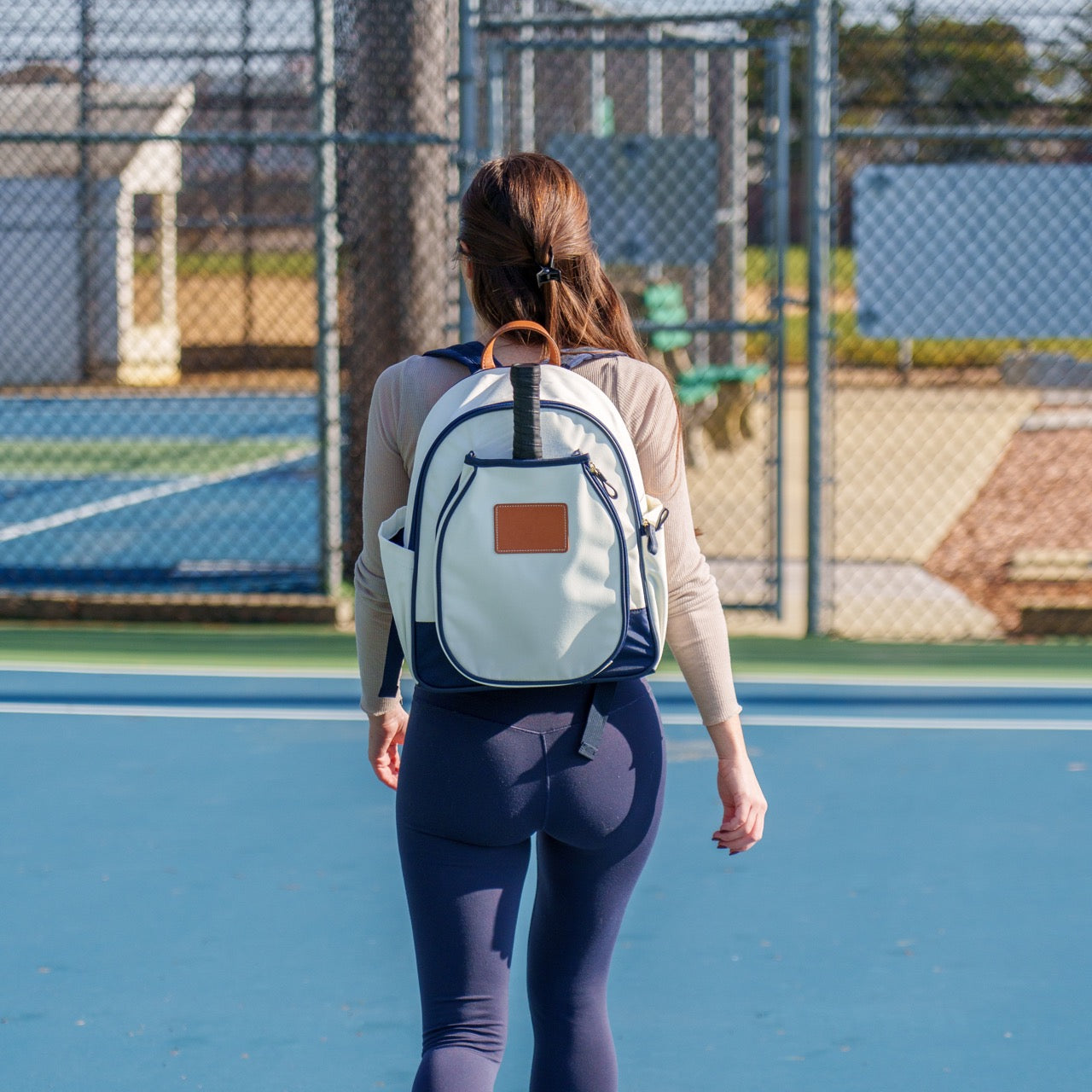 Woman walking to a pickleball court wearing a white and navy backpack with a pickleball paddle in it