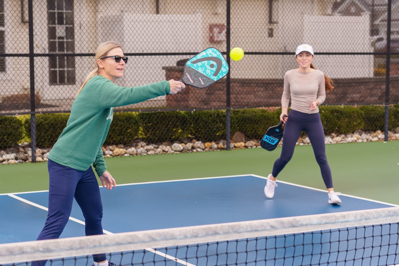 Two woman playing doubles pickleball with one currently hitting the ball