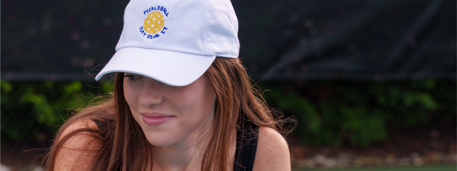 Stylish model leaning on the net of a pickleball court with a white hat that says Pickleball Just Dink It.