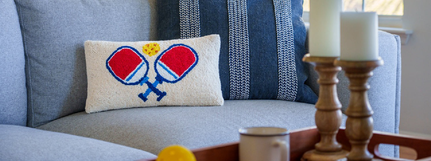 Pickleball themed hook pillow sitting on a couch in a living room with candles and a coffee mug.