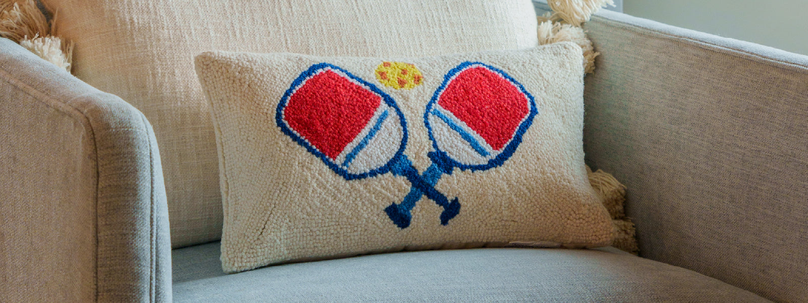 Stylish hook pillow with two pickleball paddles and a ball on the front sits on a chair in a living room.