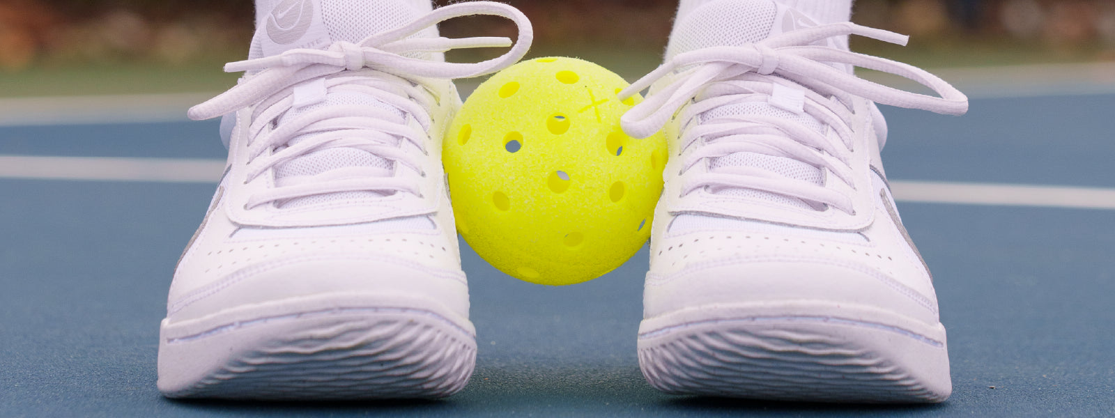 Close up of a pickleball players court shoes with a pickleball sitting in between their feet a few inches off the ground.