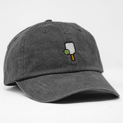 Side of washed black baseball hat that has an embroidered pickleball paddle and ball on it