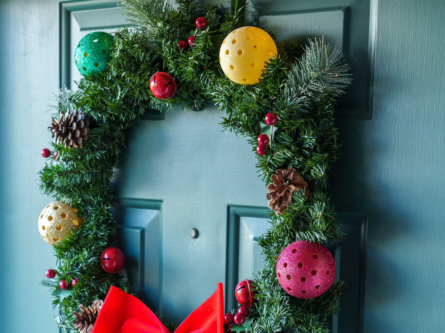 A Christmas wreath on a green front door adorned by sparkly pickleball Christmas ornaments and a red bow.