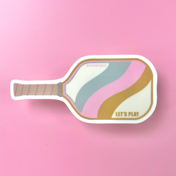 Sticker in the shape of a pickleball paddle that says Let's Play on it on a pink background