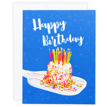 Colorful pickleball themed birthday card with a birthday cake with candles sitting on a pickleball paddle and big bold text that reads Happy Birthday laying flat on a white background.