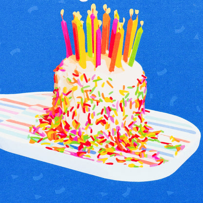 Close up of a colorful pickleball themed birthday card with a birthday cake with candles sitting on a pickleball paddle and big bold text that reads Happy Birthday laying flat on a white background.
