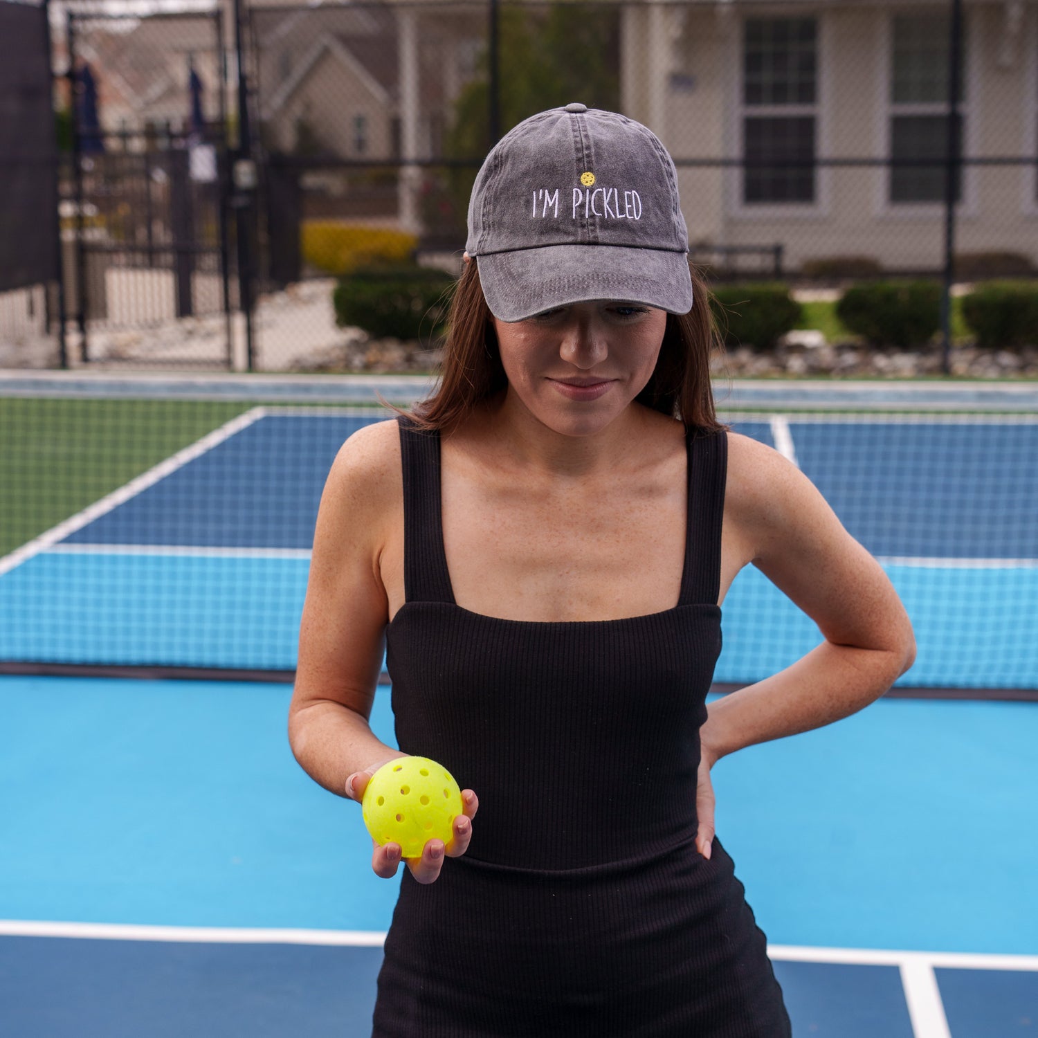 Woman standing on a pickleball court holding a pickleball and wearing a black embroidered hat that says &quot;I&