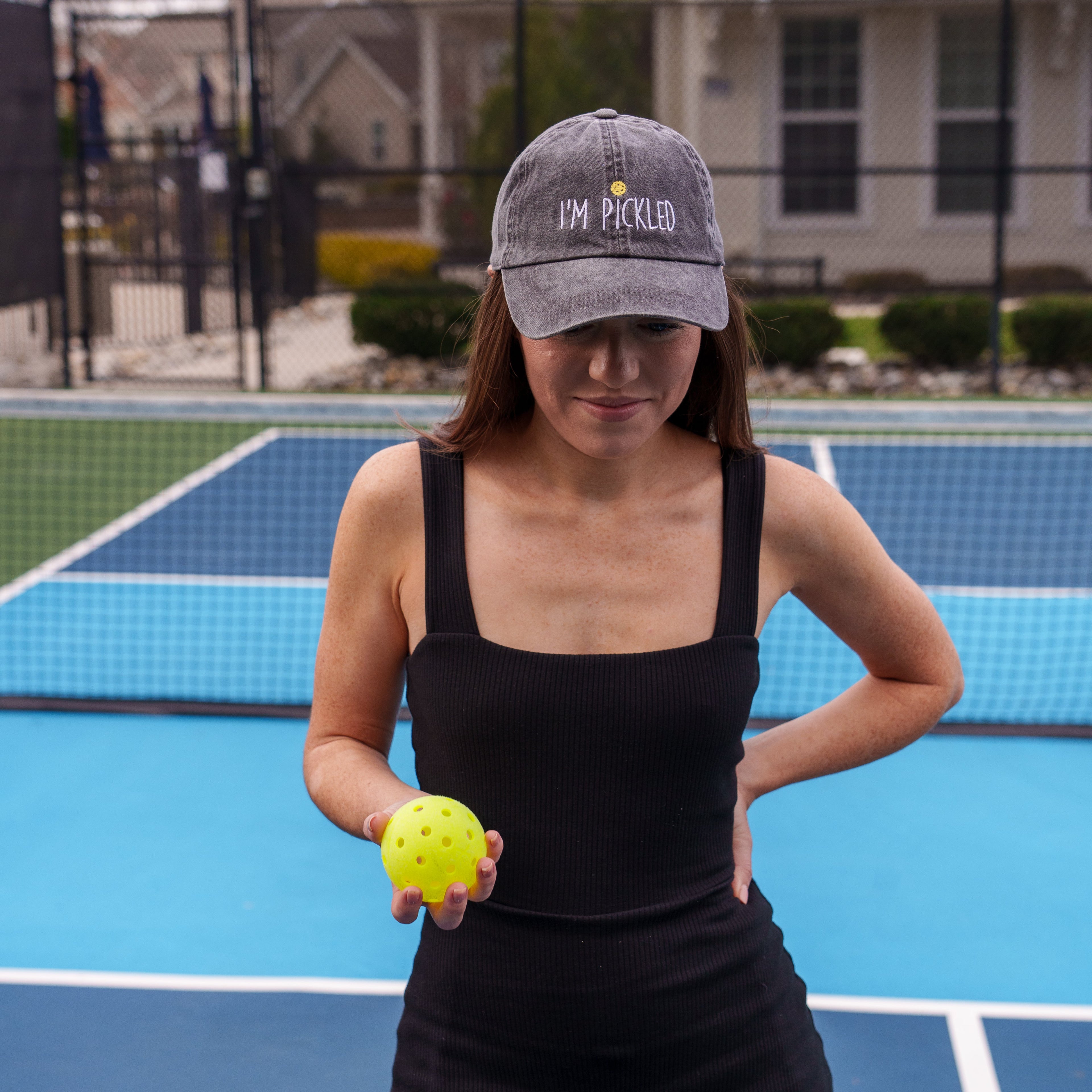 Woman standing on a pickleball court holding a pickleball and wearing a black embroidered hat that says &quot;I&