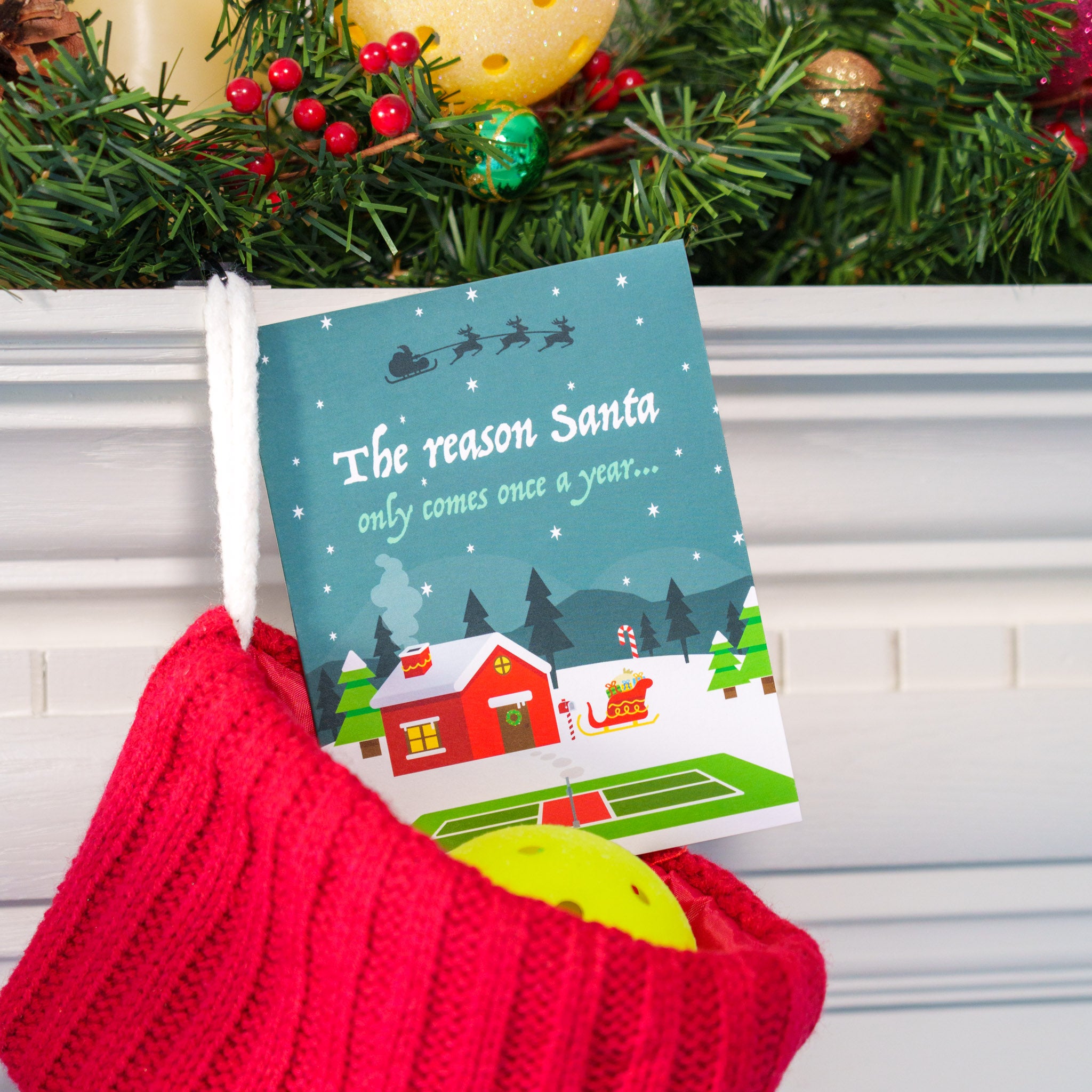 A christmas card that says &quot;The reason Santa only comes once a year...&quot; and has a pickleball court on it. The card is sitting in a stocking hanging on a mantle.