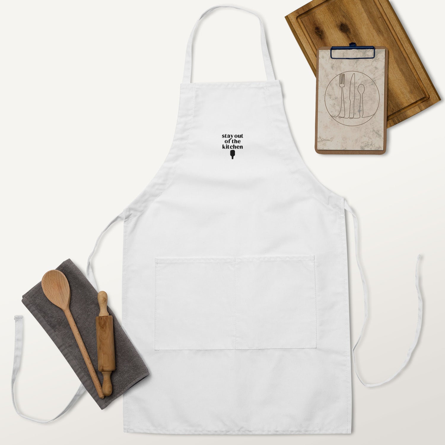 Embroidered white apron that says &quot;stay out of the kitchen&quot; and has a pickleball paddle on it with a cutting board, rolling pin and wooden spoon next to it