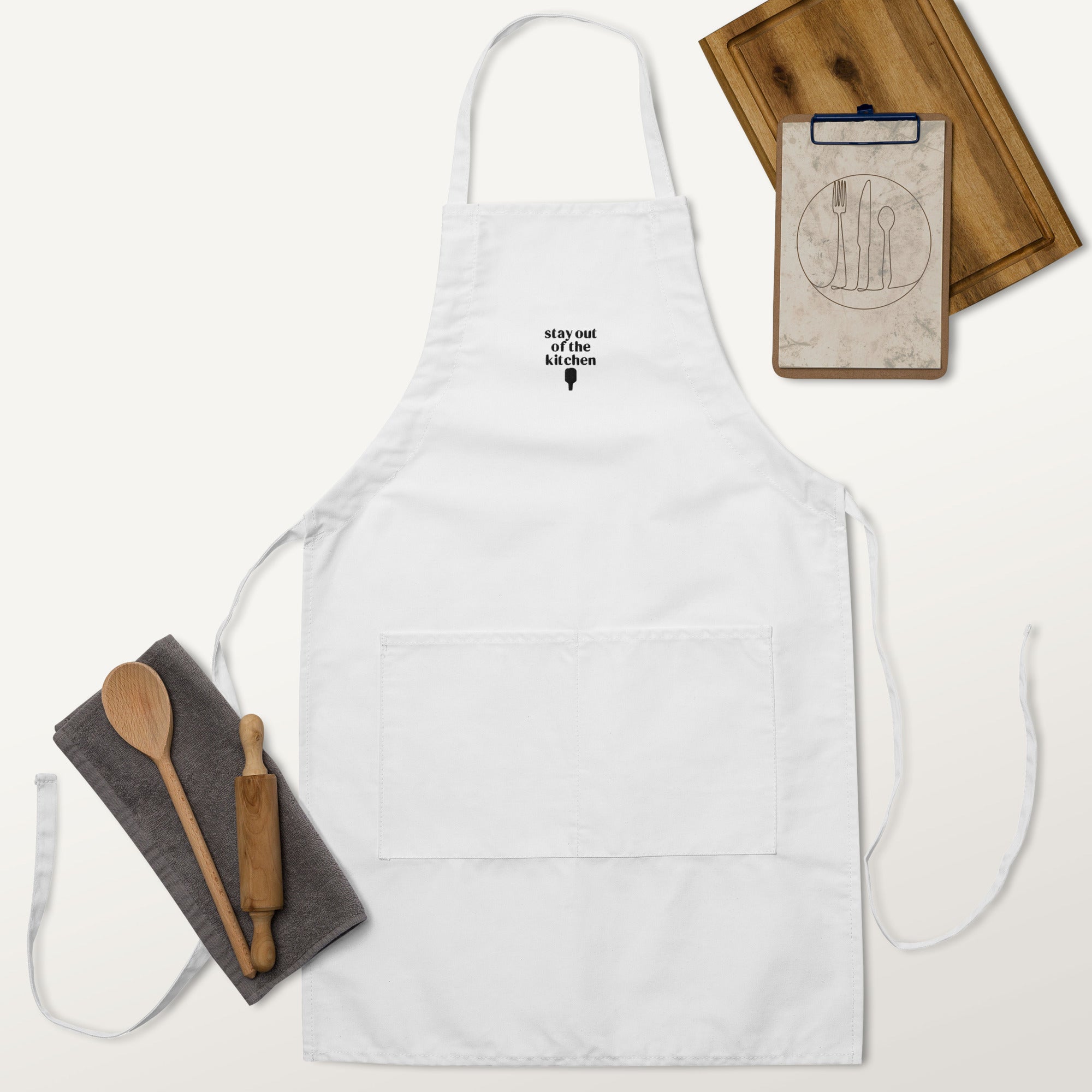 Embroidered white apron that says &quot;stay out of the kitchen&quot; and has a pickleball paddle on it with a cutting board, rolling pin and wooden spoon next to it